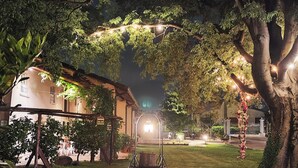 Un espacio exterior bien iluminado con una fuente, rodeado de árboles y un edificio al fondo.