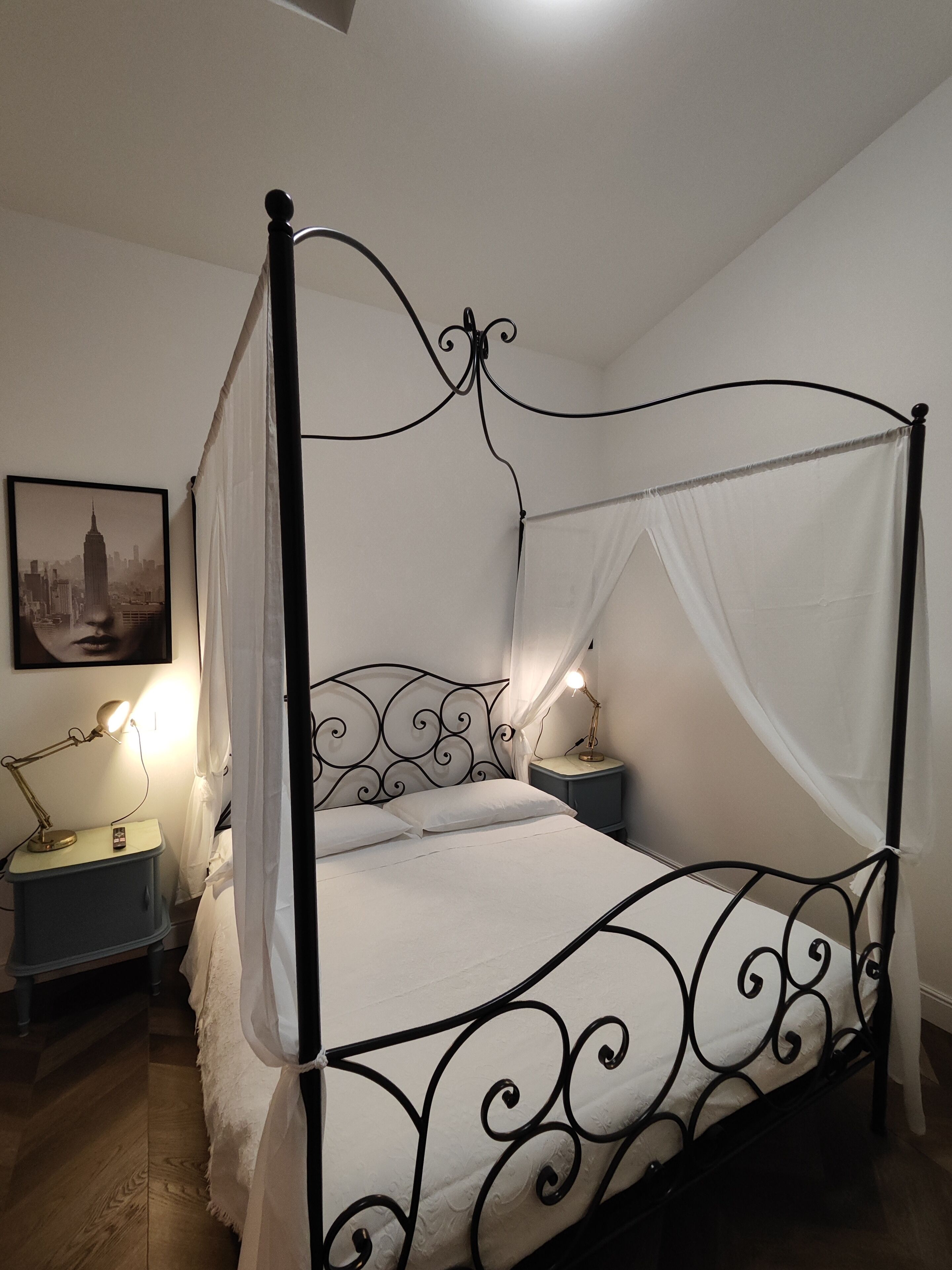 A four-poster bed with a canopy, two bedside tables with lamps, a framed picture on the wall, and a wooden floor.