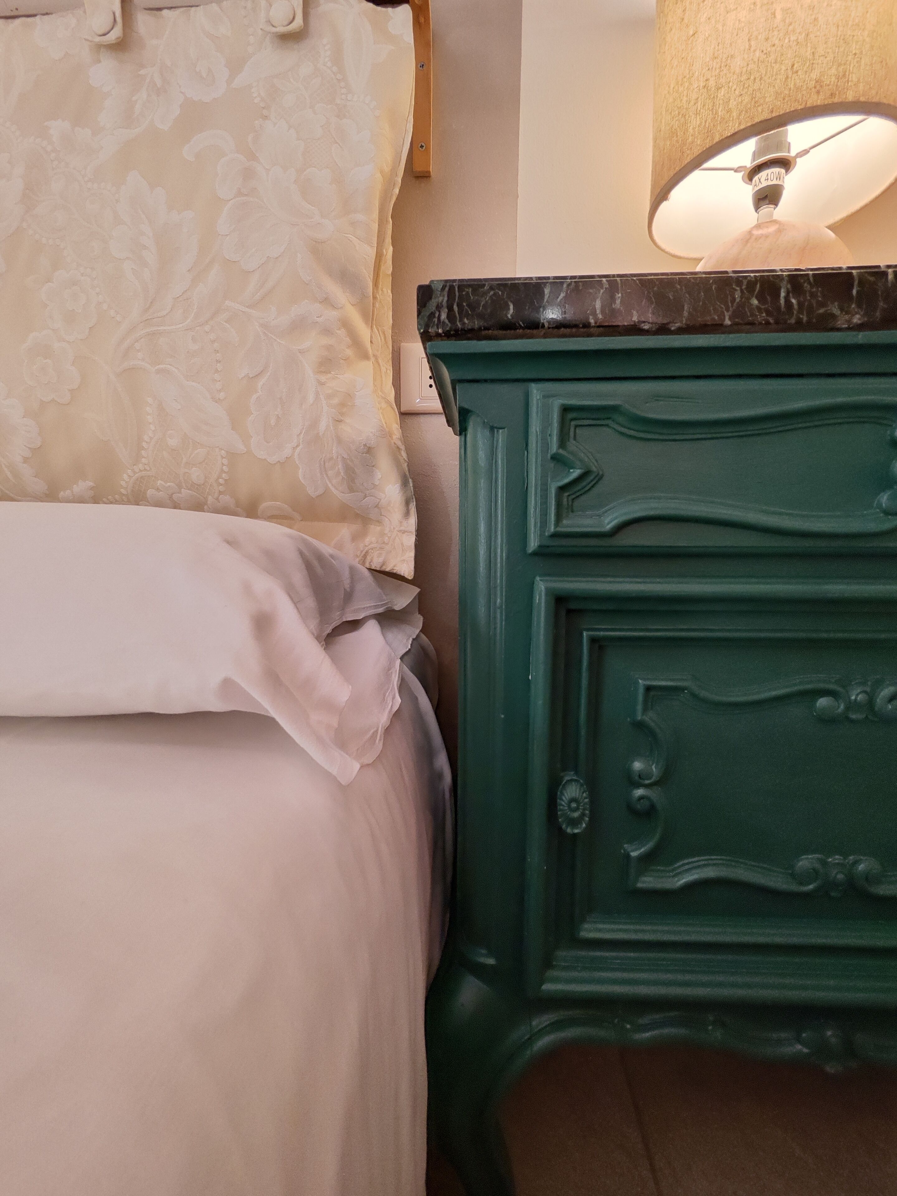 Un lit avec une literie à motifs floraux, une table de chevet verte avec des tiroirs et une lampe posée dessus.