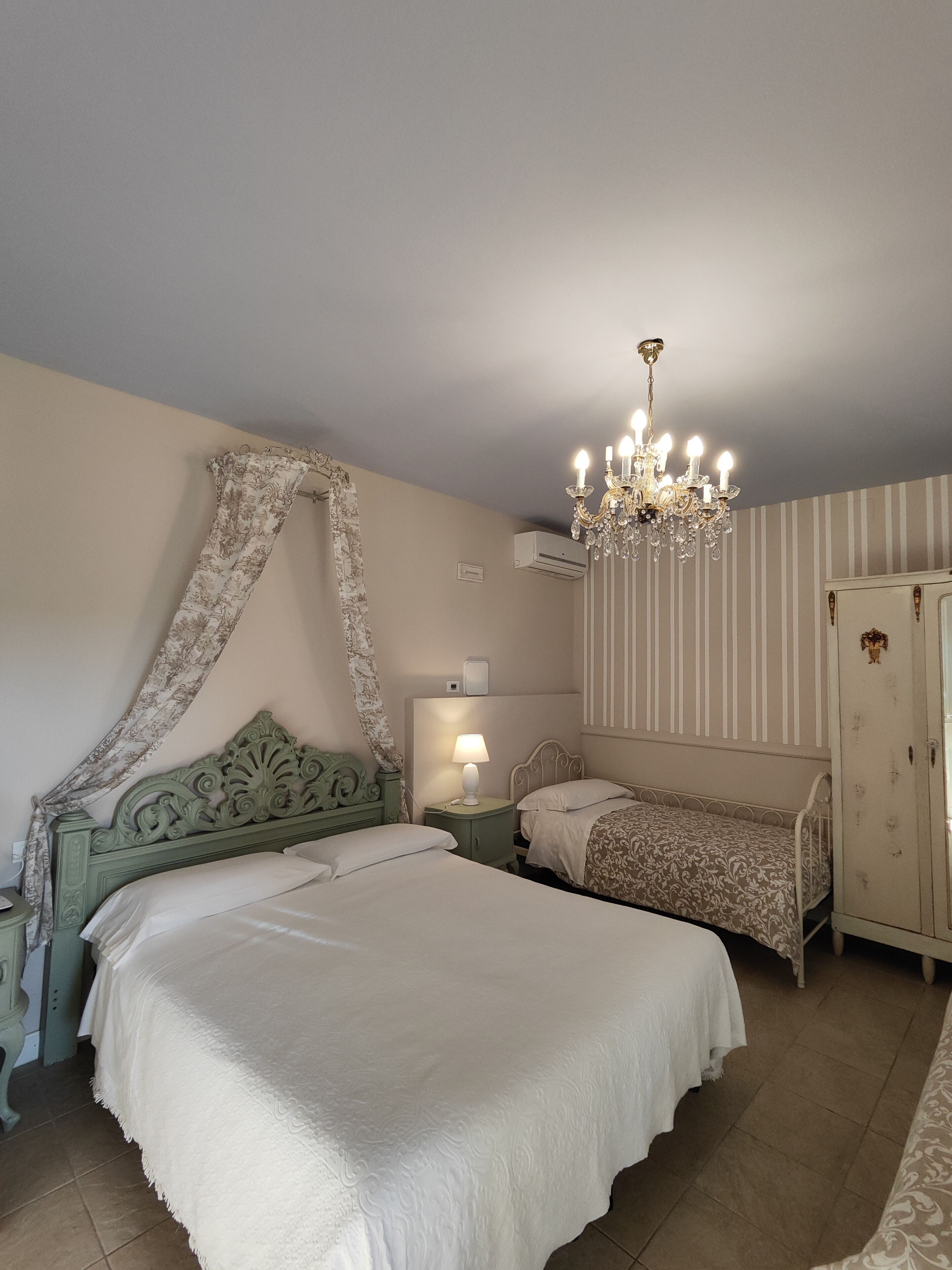 Quarto com cama de dossel, mesa de cabeceira com abajur, cômoda e lustre.
