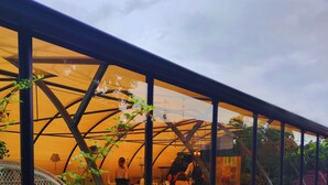 Un comedor con paredes de vidrio, mesas y sillas, un pequeño jardín con rocas y plantas, y un farol.