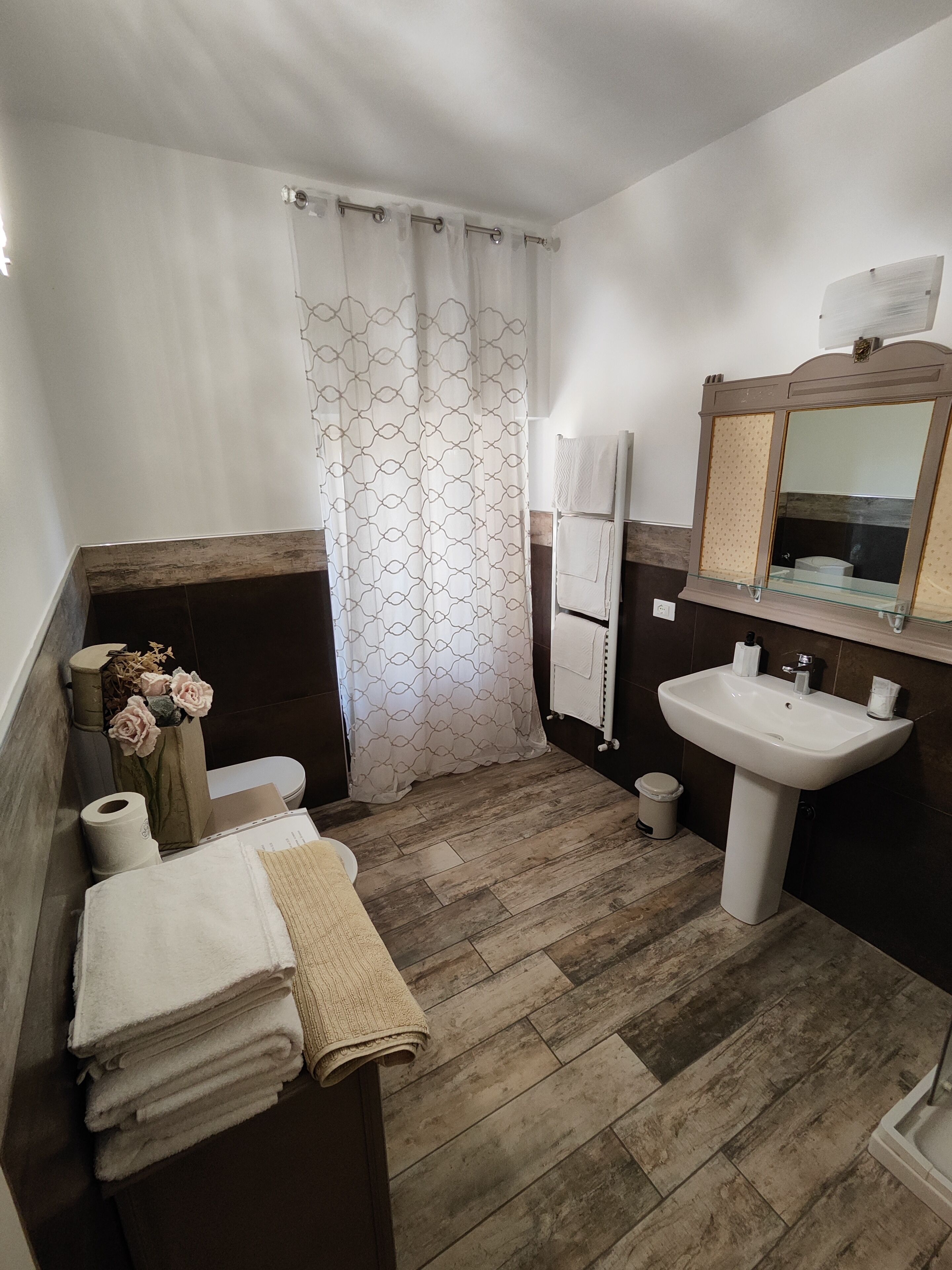 Un baño con suelo de madera, un lavamanos blanco, un espejo y una cortina de ducha con estampado geométrico.