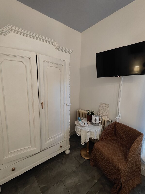 Quarto com um guarda-roupa branco, televisão na parede, uma pequena mesa com toalha rendada e uma poltrona marrom.