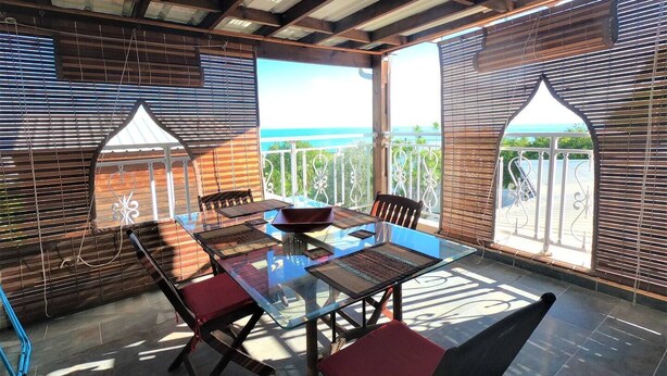 An outdoor dining area with a glass table and chairs, wooden blinds, and a view of the ocean. - LE CLOS DE VAL - ACCOMMODATION - TERRACE SEA VIEW - HEATED SWIMMING POOL  (La Possession)