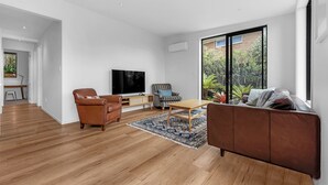 Un salon moderne avec un plancher en bois, un canapé en cuir marron, une table basse en bois, un téléviseur et une grande fenêtre donnant sur un paysage verdoyant.
