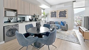 A modern kitchen with built-in appliances, a dining area with a round table and chairs, a living room with a sofa and a large window, and artwork on the wall. - 2-Bed City Escape with Mountain Views (Cape Town)