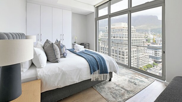A modern bedroom with a large bed, a nightstand, a lamp, and a view of a cityscape through large windows. - 2-Bed City Escape with Mountain Views (Cape Town)