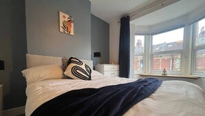 A bedroom with a bed, a nightstand, a window with a view of buildings, and a wall with a piece of art.