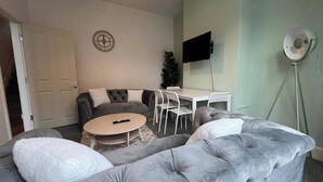 A living room with a grey sofa, a round coffee table, a white TV, and a small white table with chairs.