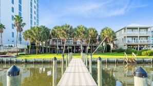 A waterfront walkway leading to a building with a balcony, surrounded by palm trees and a calm water body. - Laguna Bella B5 (Gulf Shores)
