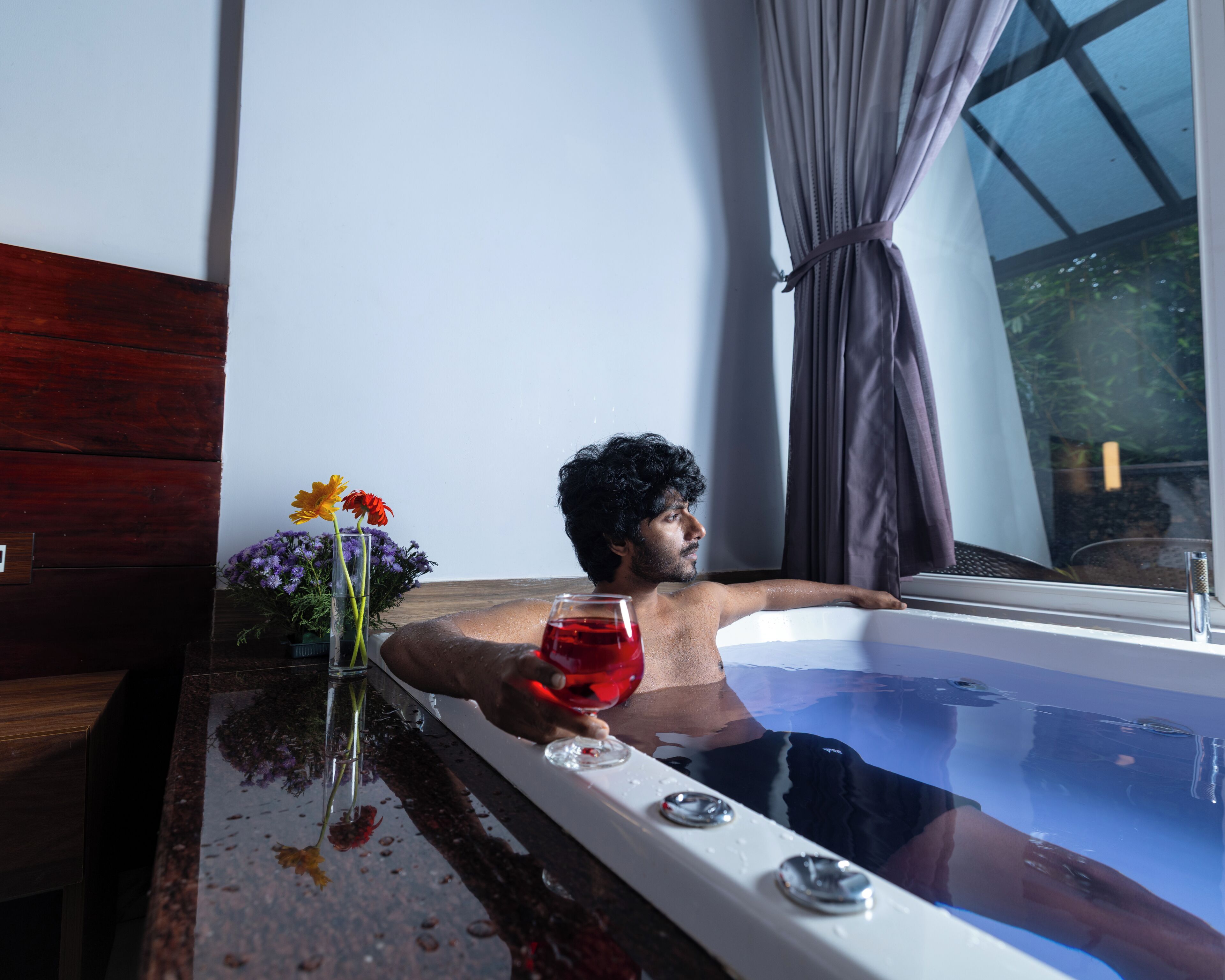 A person relaxing in a bathtub with flowers and a glass of red liquid on the side.
