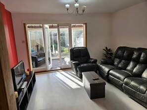 A living room with a black leather sofa, a black armchair, a coffee table, and a TV on a stand. There is a sliding glass door leading to a balcony. - Entire Treasure of Nottingham (Nottingham)