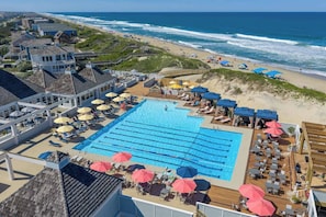 Ein großer Pool mit Liegestühlen und Sonnenschirmen, im Hintergrund ein Strand, und ein Gebäude mit einem Balkon mit Blick auf den Pool.