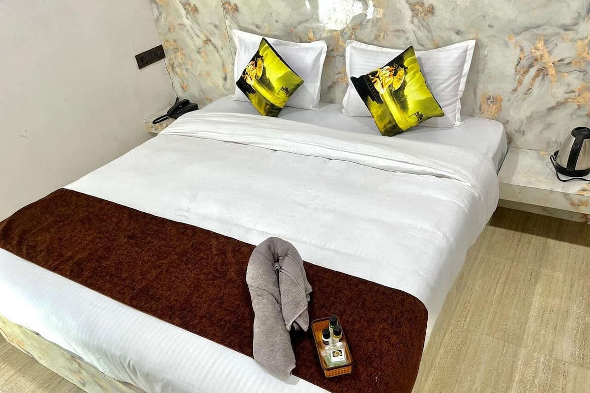 Una cama bien hecha con sábanas blancas y dos almohadones decorativos, una toalla marrón y una botella de loción en la mesita.
