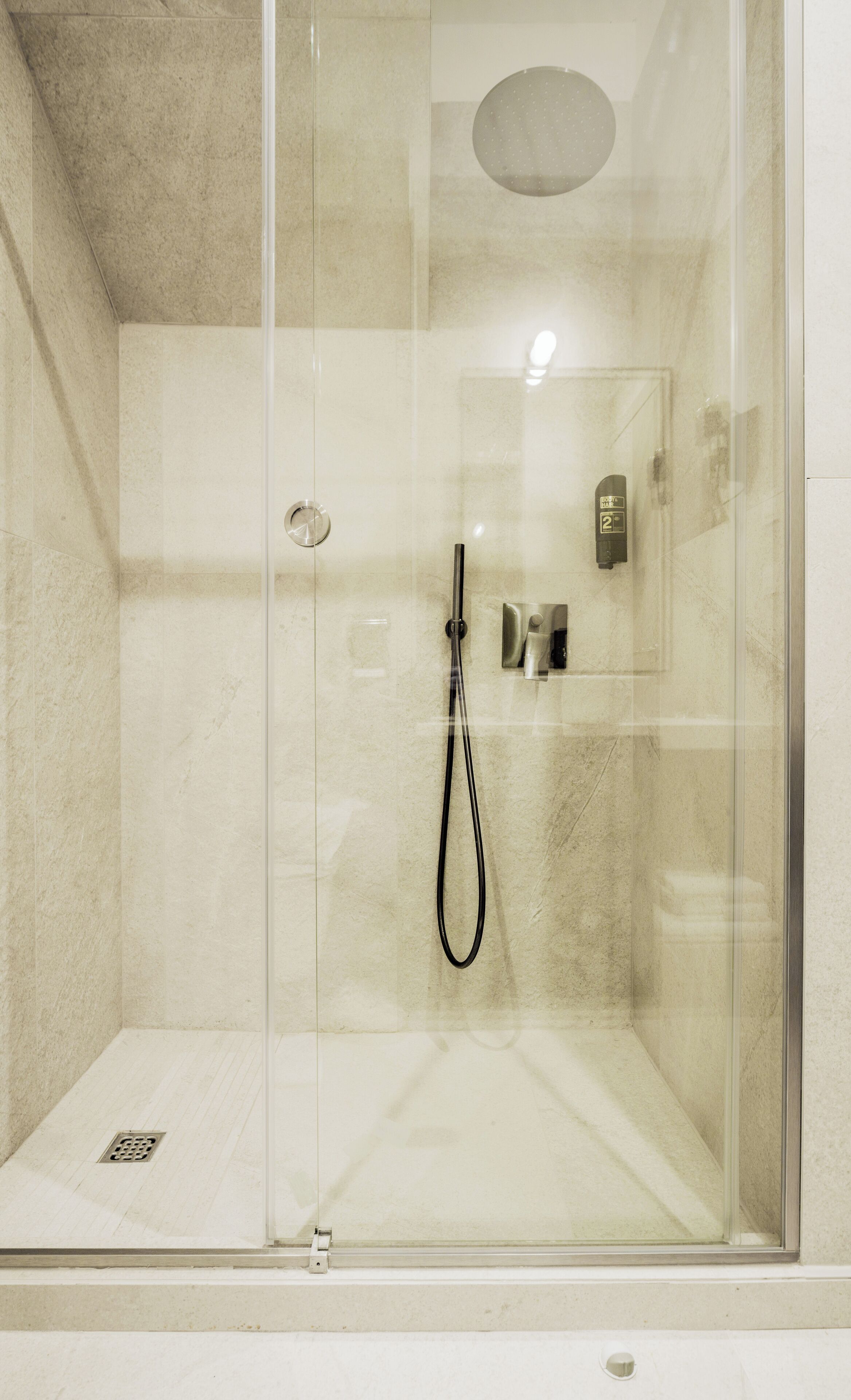 Une cabine de douche fermée par des parois en verre, dotée d’un pommeau de douche à main, d’un panneau de commande et d’un drain.