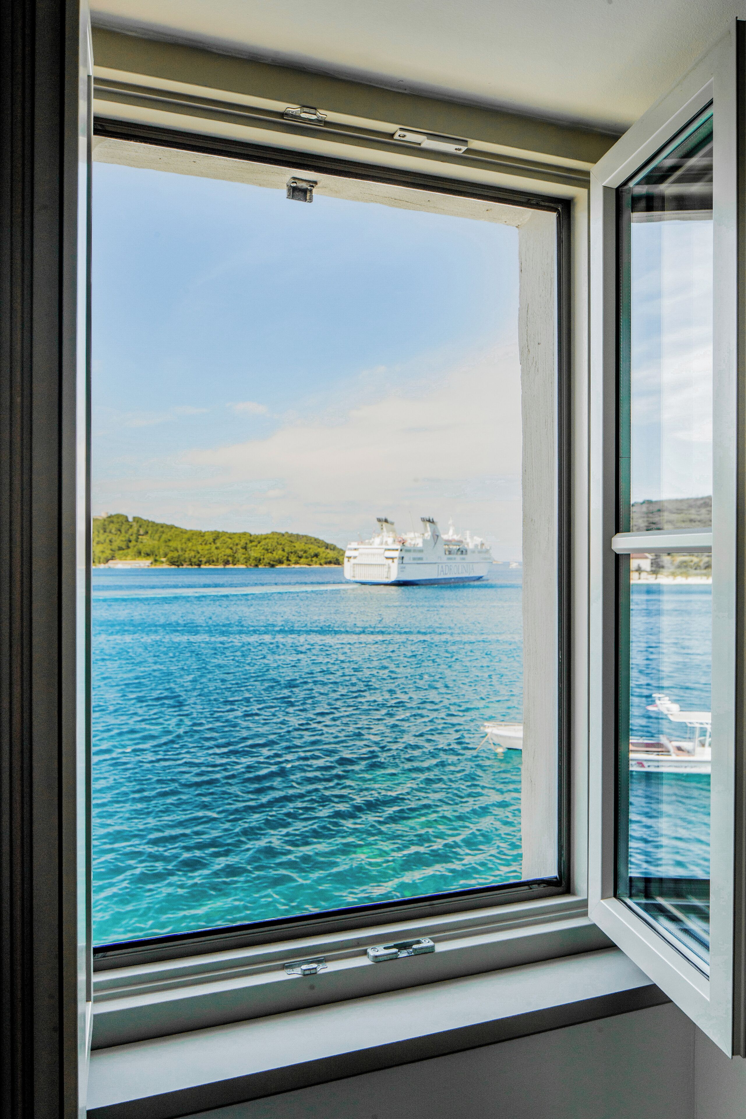 Een raam met uitzicht op een groot cruiseschip op het water, een heldere lucht en een verafgelegen kustlijn met begroeiing.