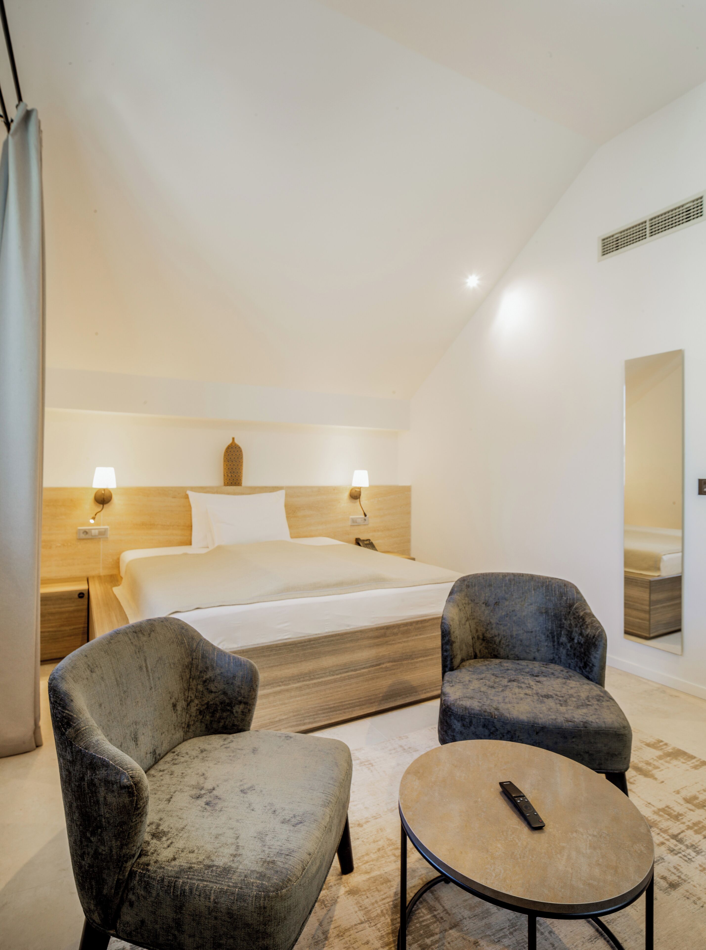 Ein modernes Hotelzimmer mit einem großen Bett, zwei Sesseln, einem kleinen runden Tisch und einem Spiegel.