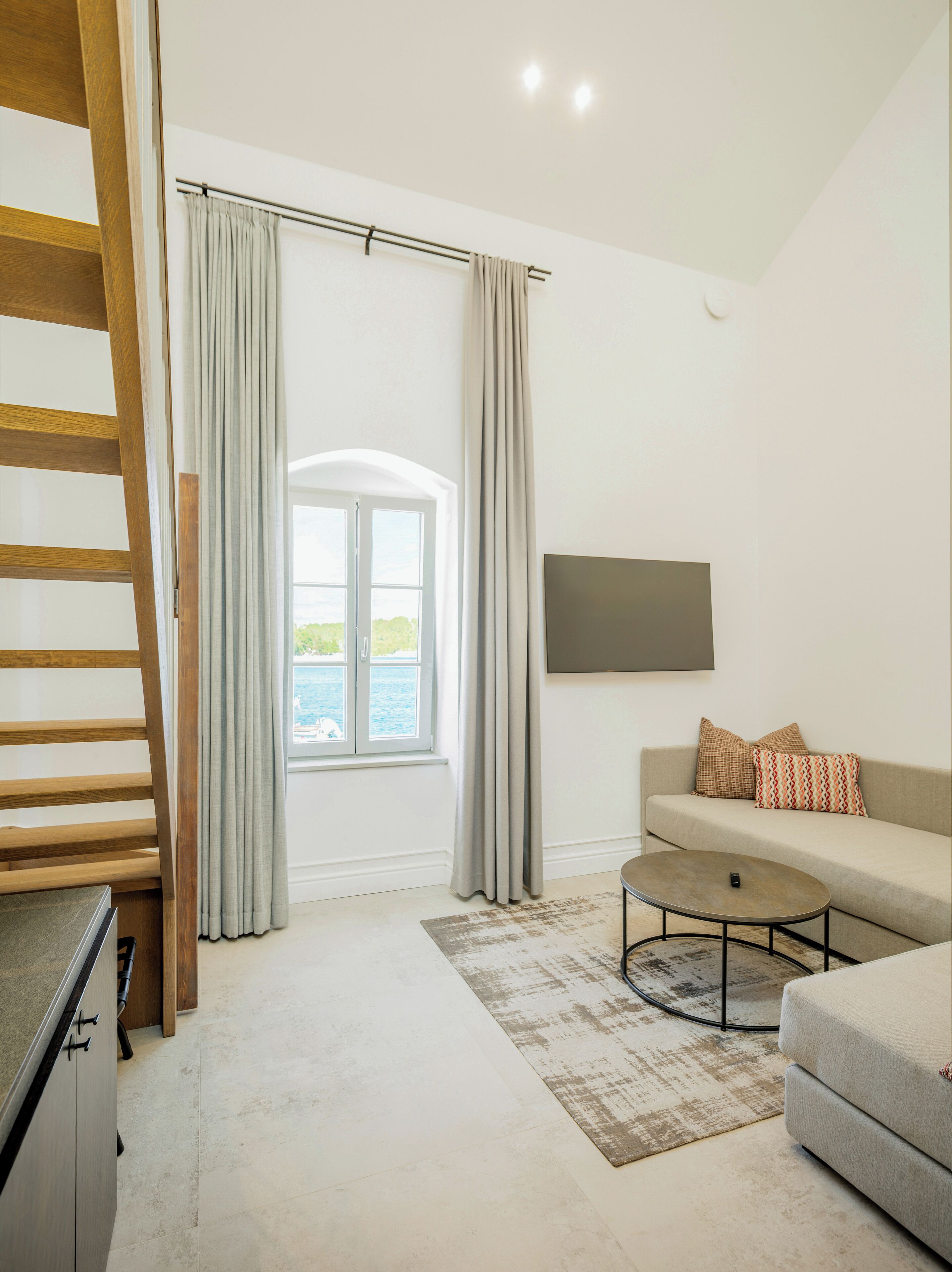 A modern living room with a sofa, a coffee table, and a TV mounted on the wall. There is a window with curtains, a view of the outdoors, and a wooden staircase.