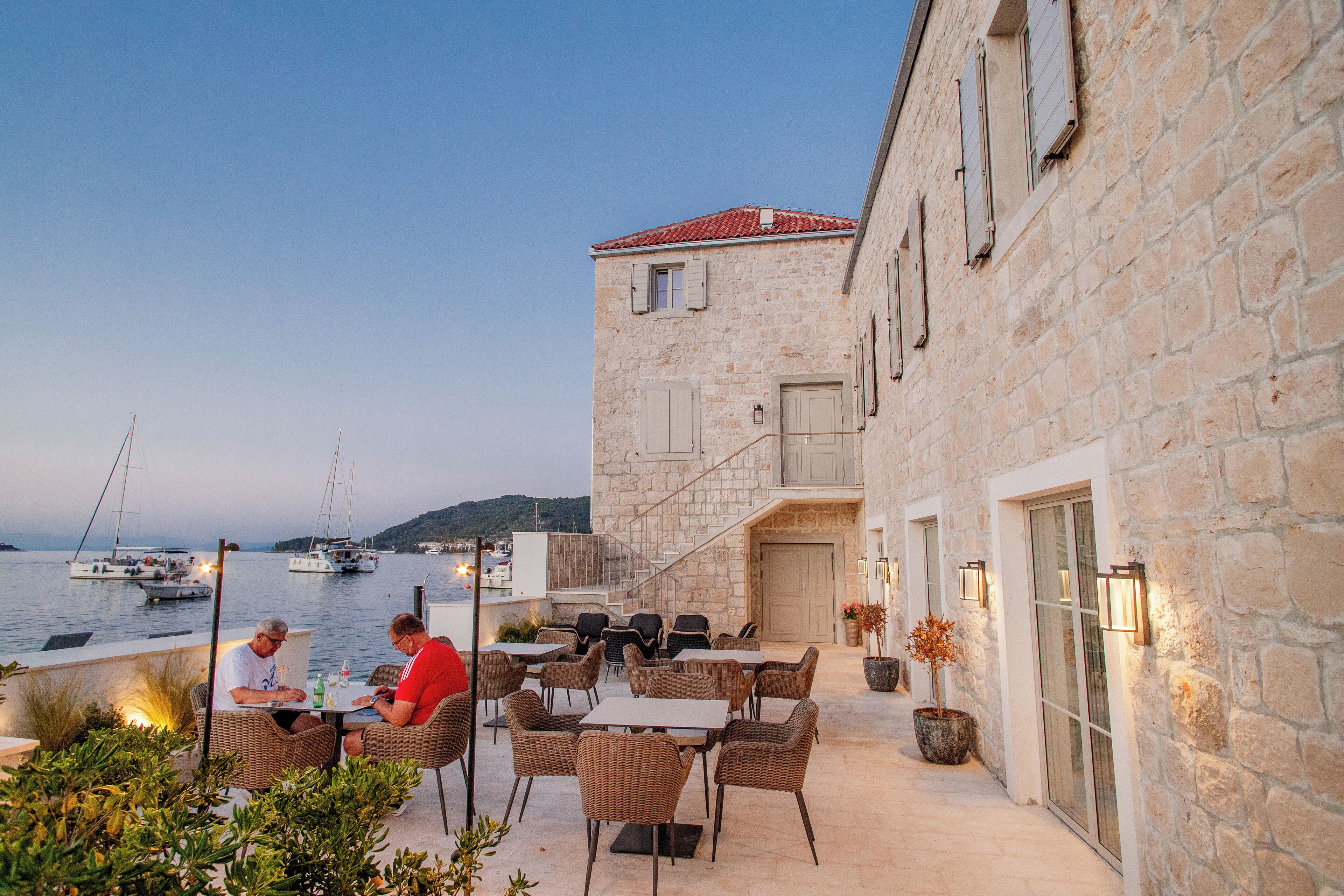 A stone building with outdoor seating by the water, featuring a staircase and a view of boats.