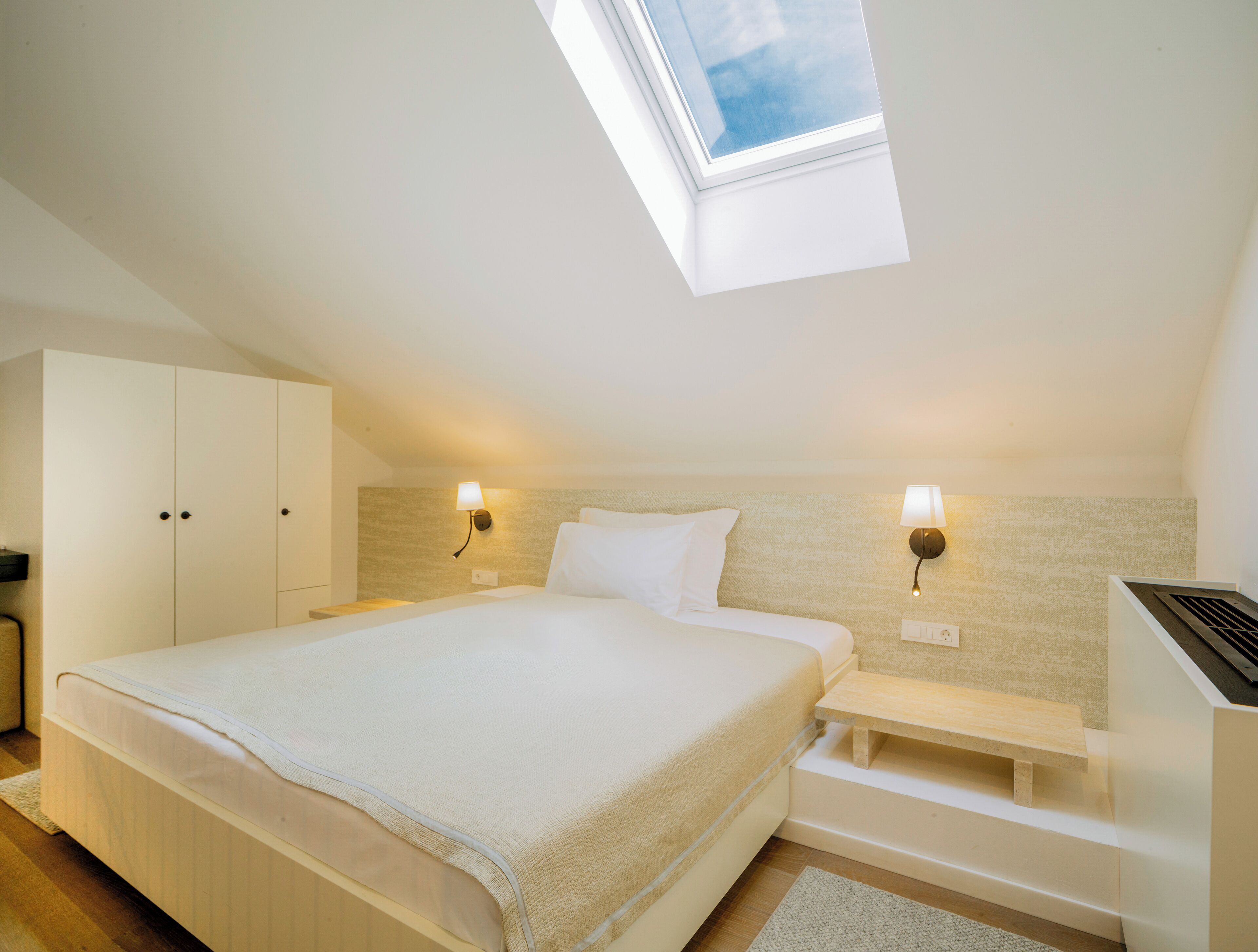 A bedroom with a skylight, a bed with white bedding, two bedside lamps, a wooden bench, and a wardrobe.