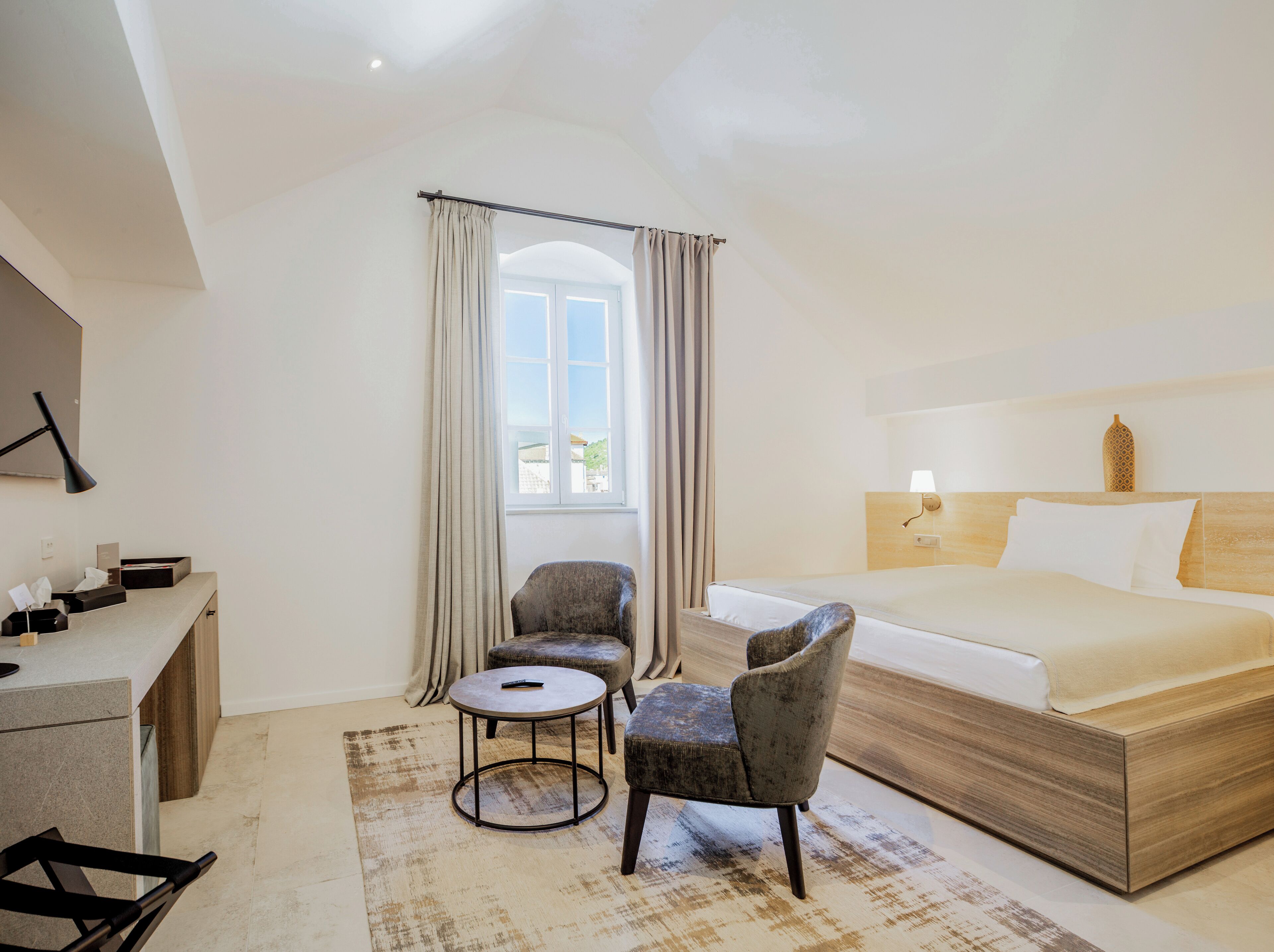 A hotel room with a bed, two armchairs, a small round table, a desk with a lamp, and a window with curtains.