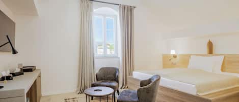 Habitación de hotel con una cama, dos sillones, una mesita redonda, un escritorio con lámpara y un ventanal con cortinas.