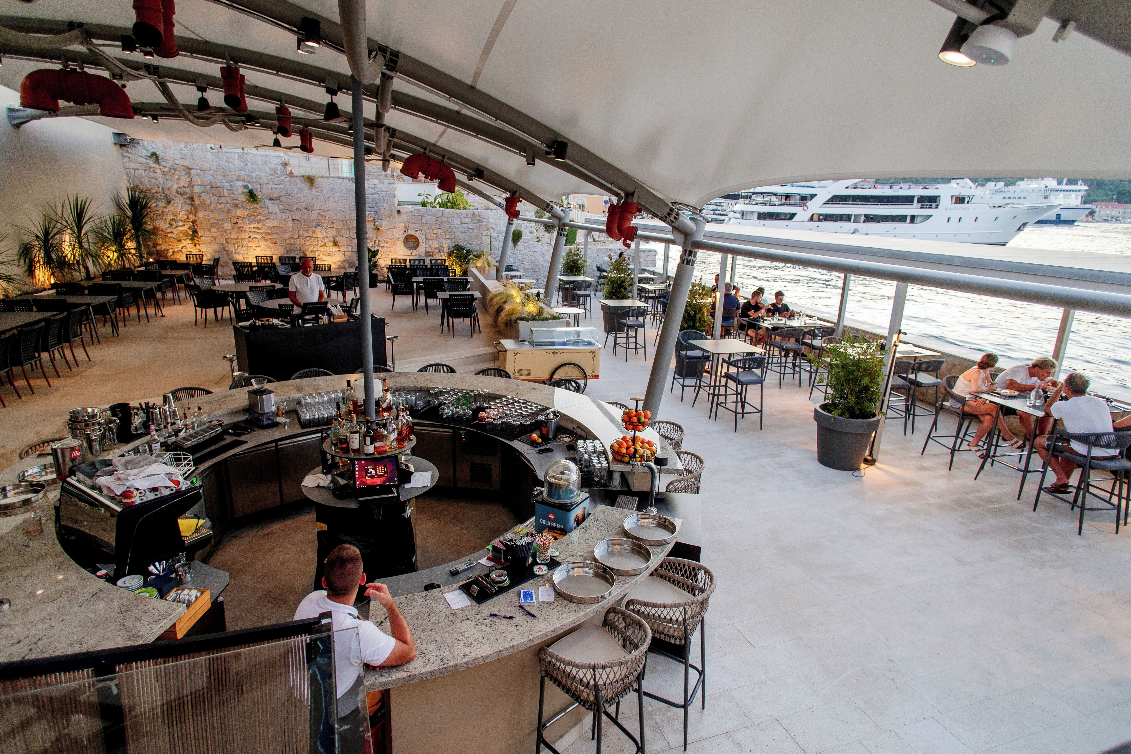 En restaurant med barområde, borde og stole, med udsigt over vandet hvor en sejlbåd kan ses i baggrunden.