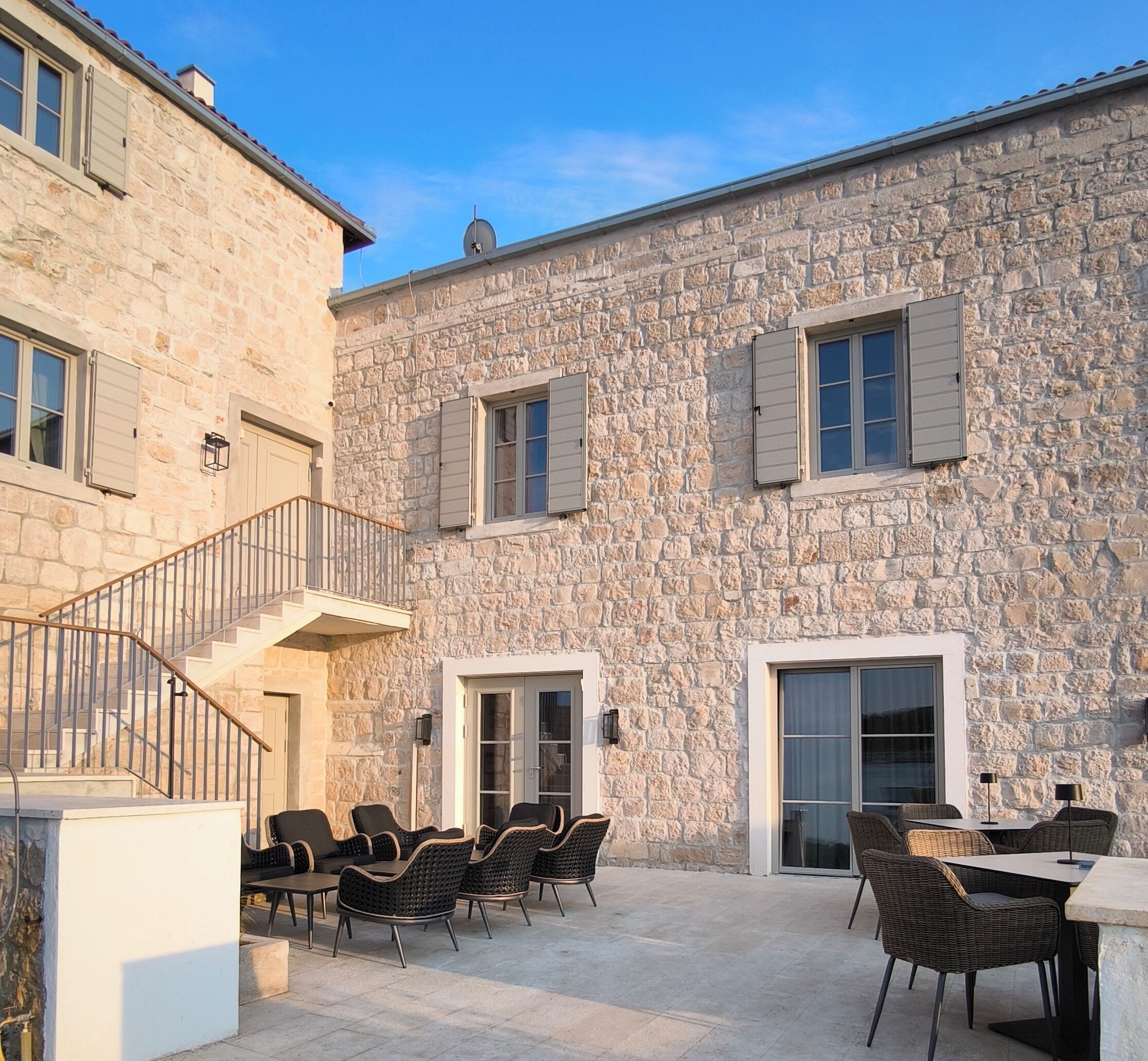 Un bâtiment en pierre avec un escalier menant à un niveau supérieur, un espace extérieur aménagé avec des chaises et des tables en osier, et un ciel bleu dégagé.
