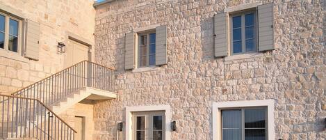 Un bâtiment en pierre avec un escalier menant à un niveau supérieur, un espace extérieur aménagé avec des chaises et des tables en osier, et un ciel bleu dégagé.