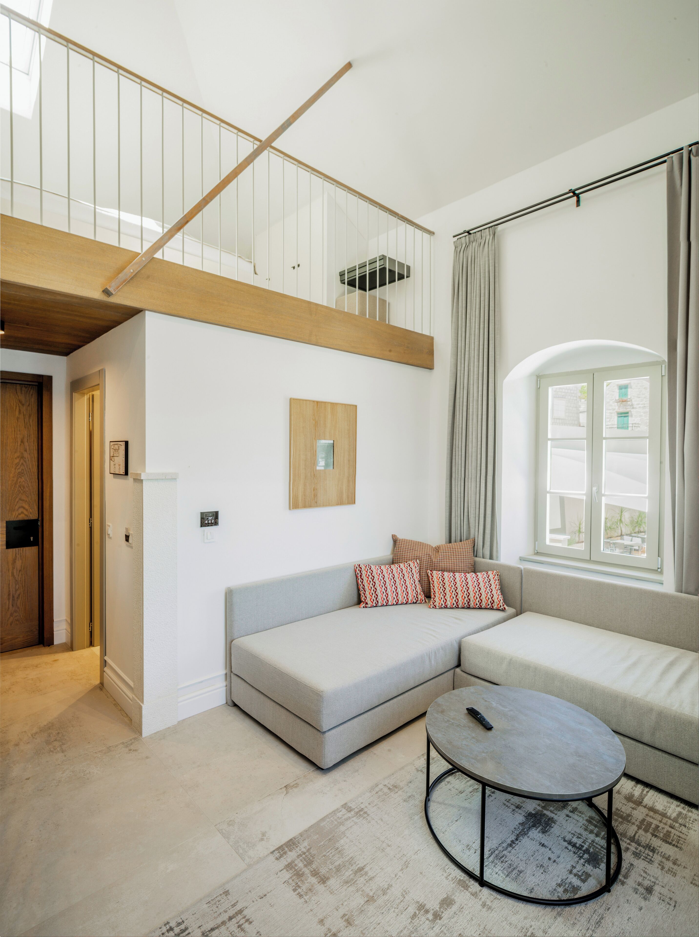 Een moderne woonkamer met een lage balkenplafond, een houten trap, een grijze bank met gekleurde kussens, een ronde salontafel en een groot raam met gordijnen.