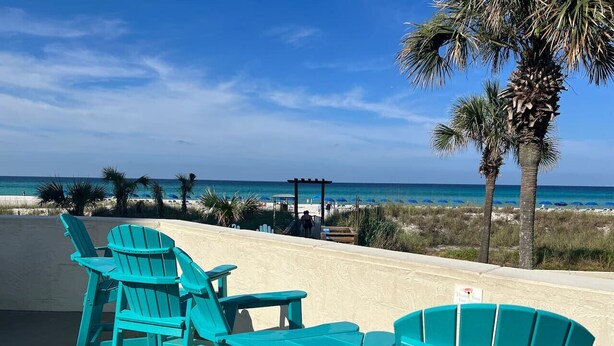 Un área de descanso frente a la playa con sillas Adirondack turquesa y una mesa de madera, con vista al océano.