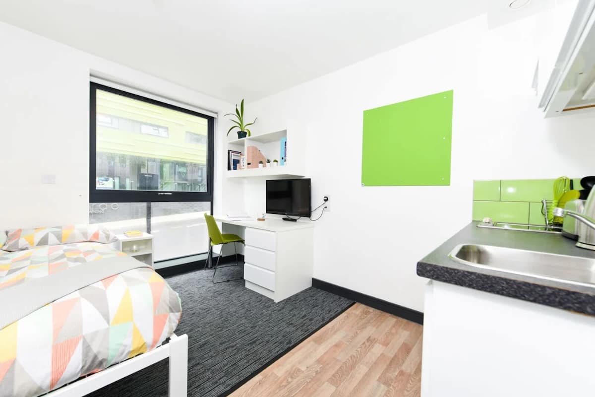A compact hotel room with a bed, a desk with a computer, a small kitchenette, and a window with a view of buildings.