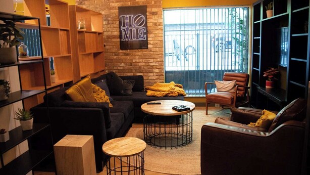 A cozy living room with a dark sofa, a wooden coffee table, and a leather armchair. There are shelves with plants and a 'HOME' sign on the wall. - Studios and Ensuites at St James Point in Newcastle (Newcastle-upon-Tyne)