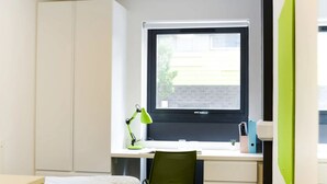 A compact bedroom with a single bed, a desk with a computer, a green chair, and a window with a view of a building. - Studios and Ensuites at St James Point in Newcastle (Newcastle-upon-Tyne)