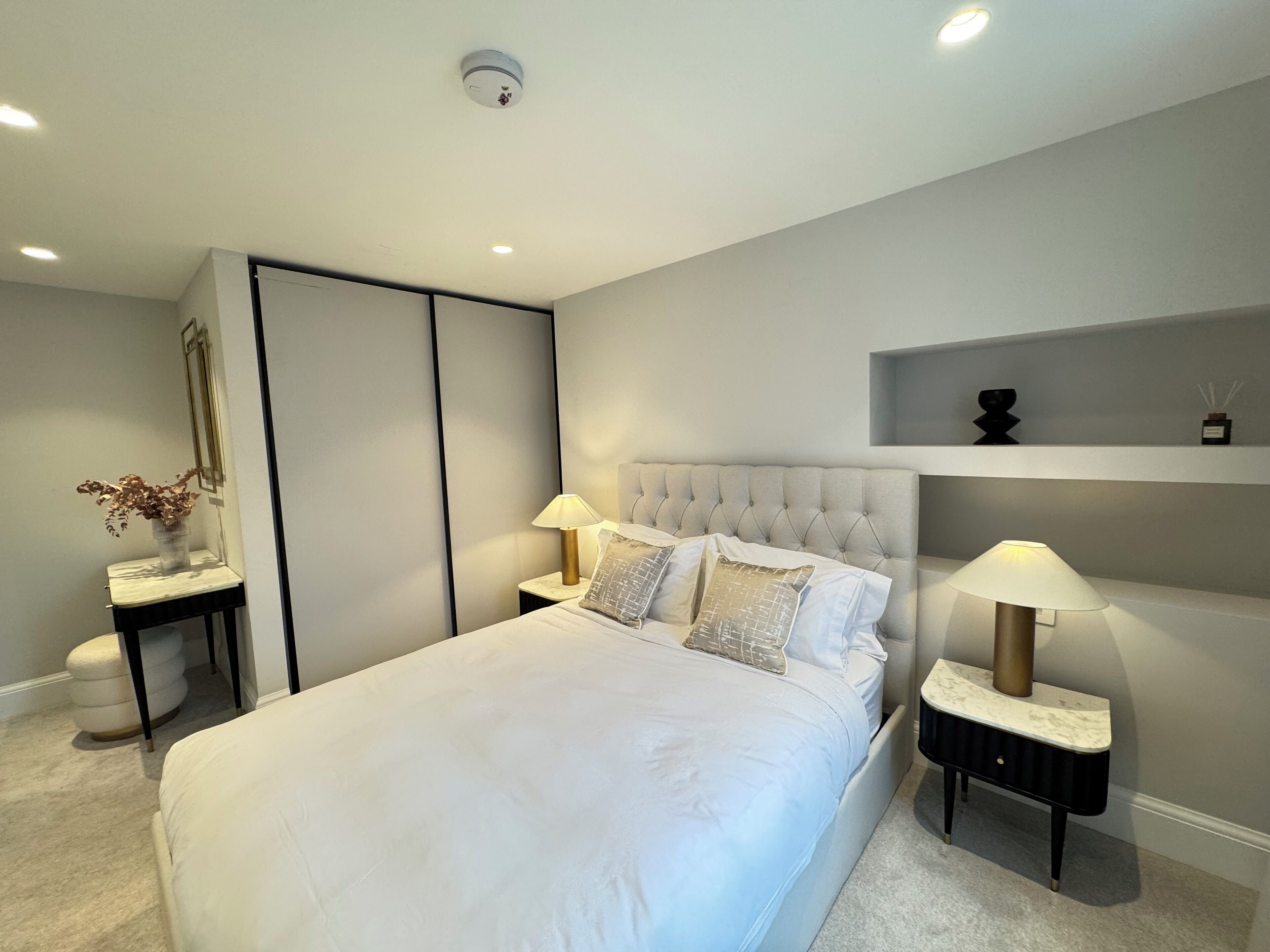 Ein modernes Schlafzimmer mit einem großen Bett, zwei Nachttischen mit Lampen, einem Einbauschrank und einem Regal mit dekorativen Accessoires.