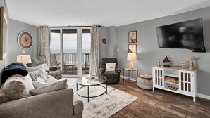 A living room with a sofa, armchairs, a coffee table, a TV, and a bookshelf. - Stunning 2 bedroom, oceanfront condo (North Topsail Beach)