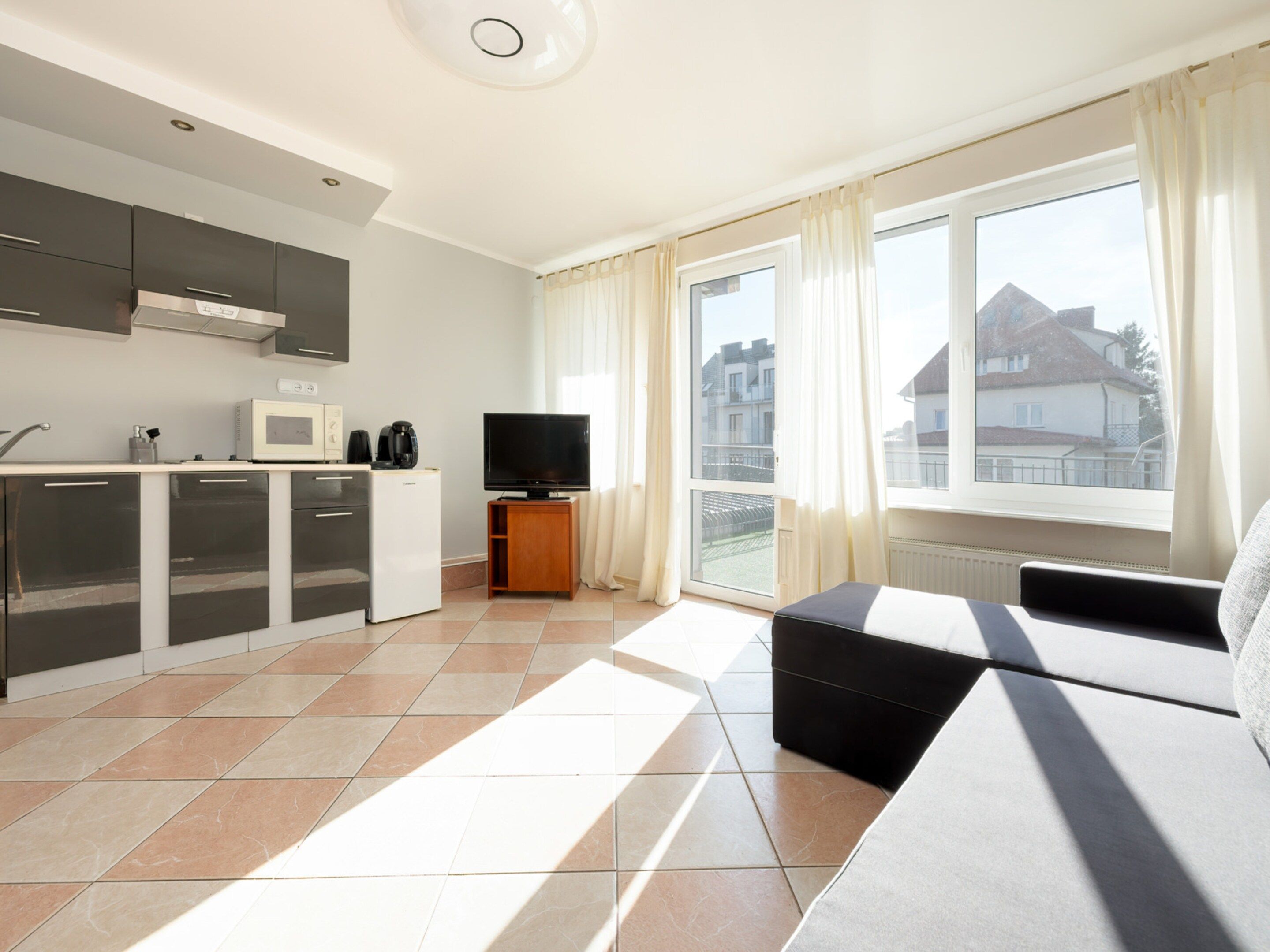 A modern apartment with a kitchen, a living area with a sofa, a TV, and a balcony with a view of buildings.