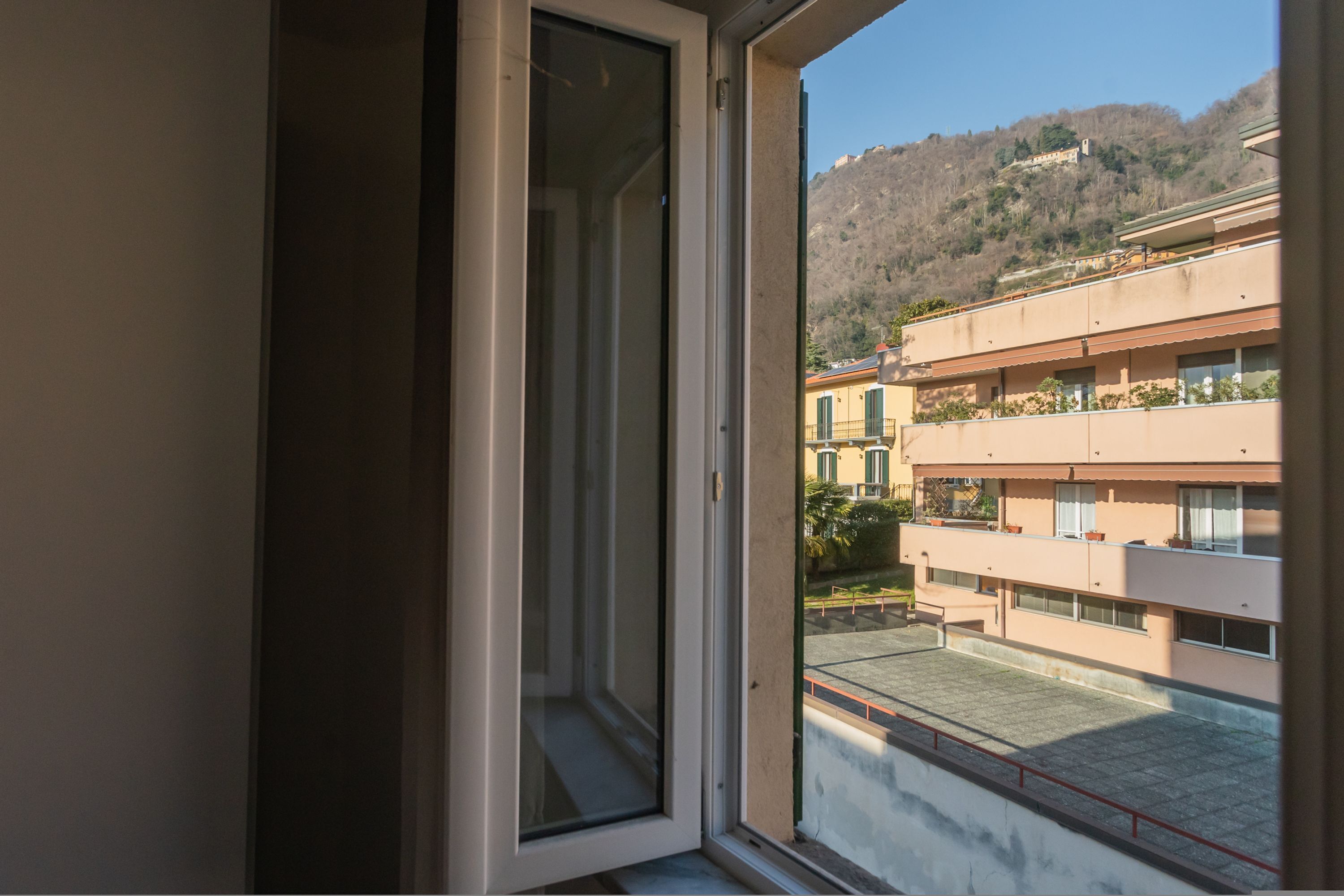 A window with a view of a multi-story building with balconies and a hilly landscape in the background.