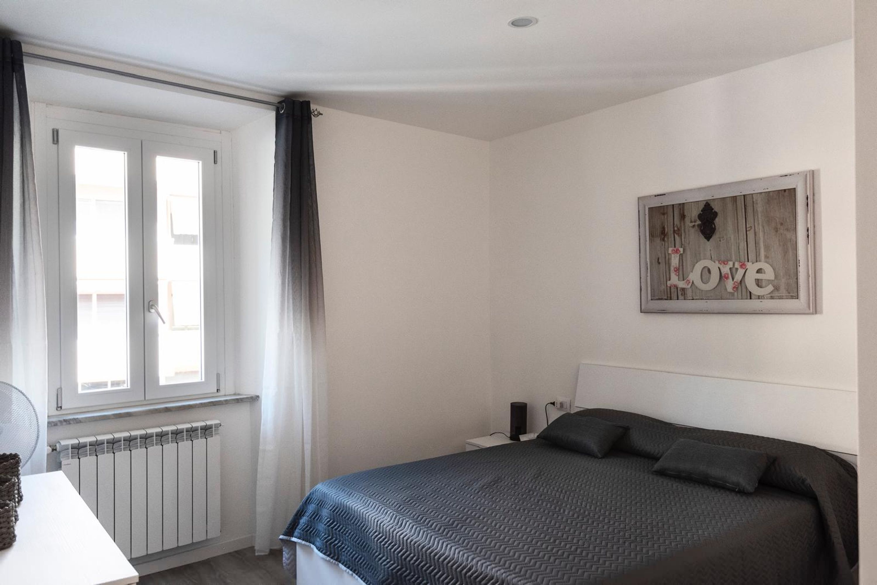 A bedroom with a bed, a radiator, a window with curtains, and a 'Love' sign on the wall.