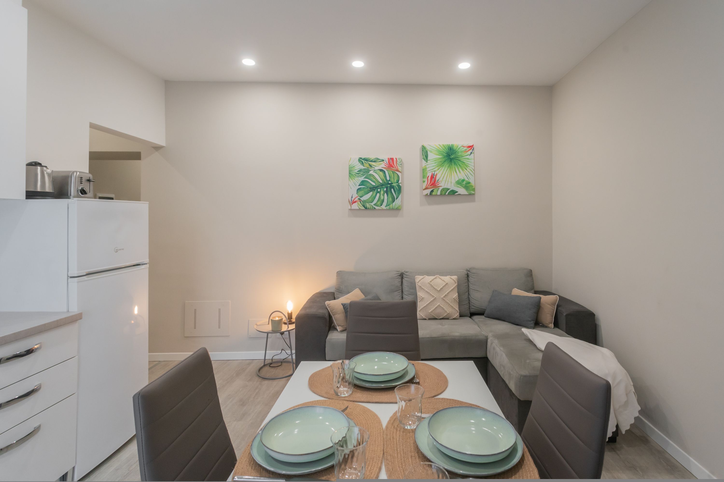 A modern kitchen with white cabinets, a built-in oven, and a coffee machine. There is a dining table with plates and glasses, and a gray sofa in the living area.