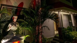 A statue of a seated figure in a garden area with palm trees and a patterned column. - Ganga Bitan Family Inn (Uluberiya)