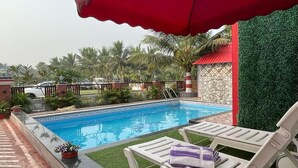 A swimming pool with lounge chairs and a red umbrella, surrounded by greenery and palm trees. - Ganga Bitan Family Inn (Uluberiya)