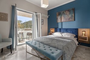 A bedroom with a large bed, a bench, a nightstand with a lamp, and a view of the outdoors. - Captain's Seafront Apartments (Lefkada)
