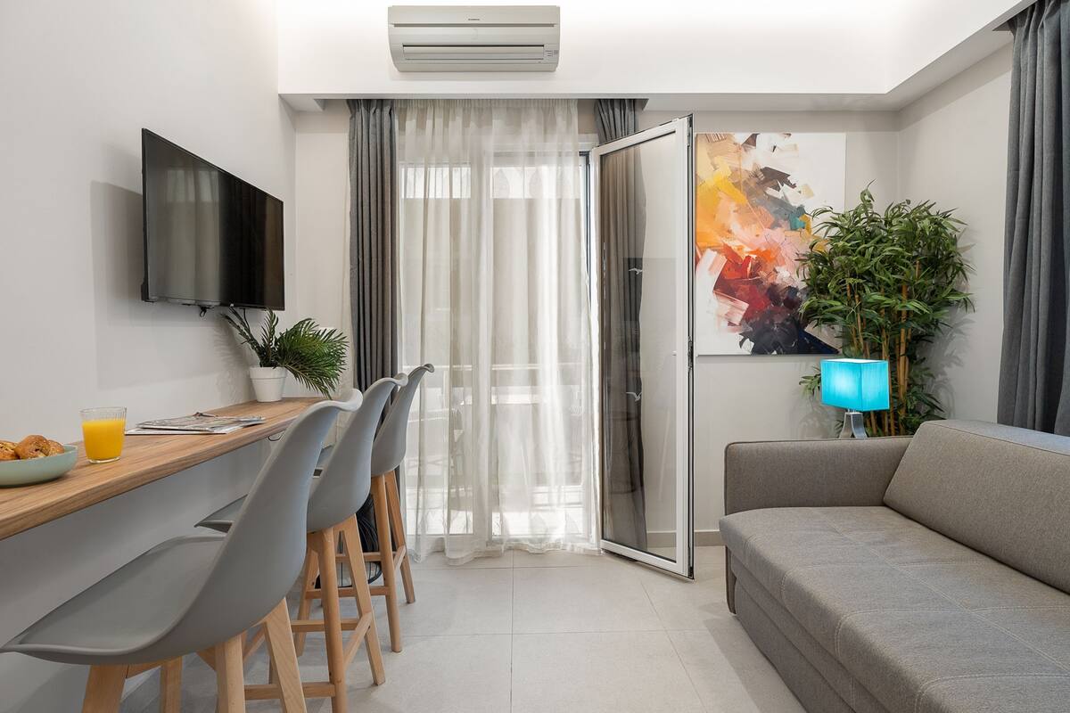 A modern living room with a grey sofa, a dining table with chairs, a flat-screen TV, and a large window with sheer curtains.