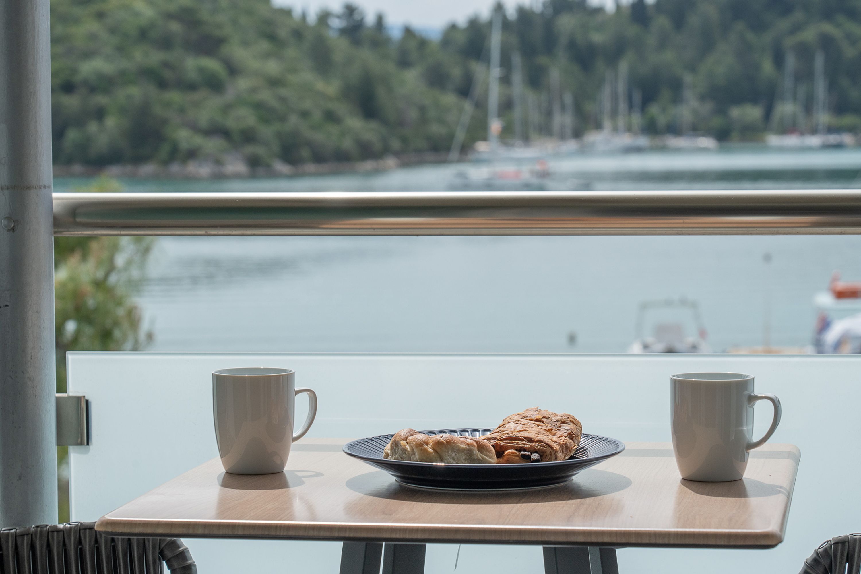 Mesa com prato de doces e duas chávenas, com vista para um corpo de água com barcos e árvores ao fundo.