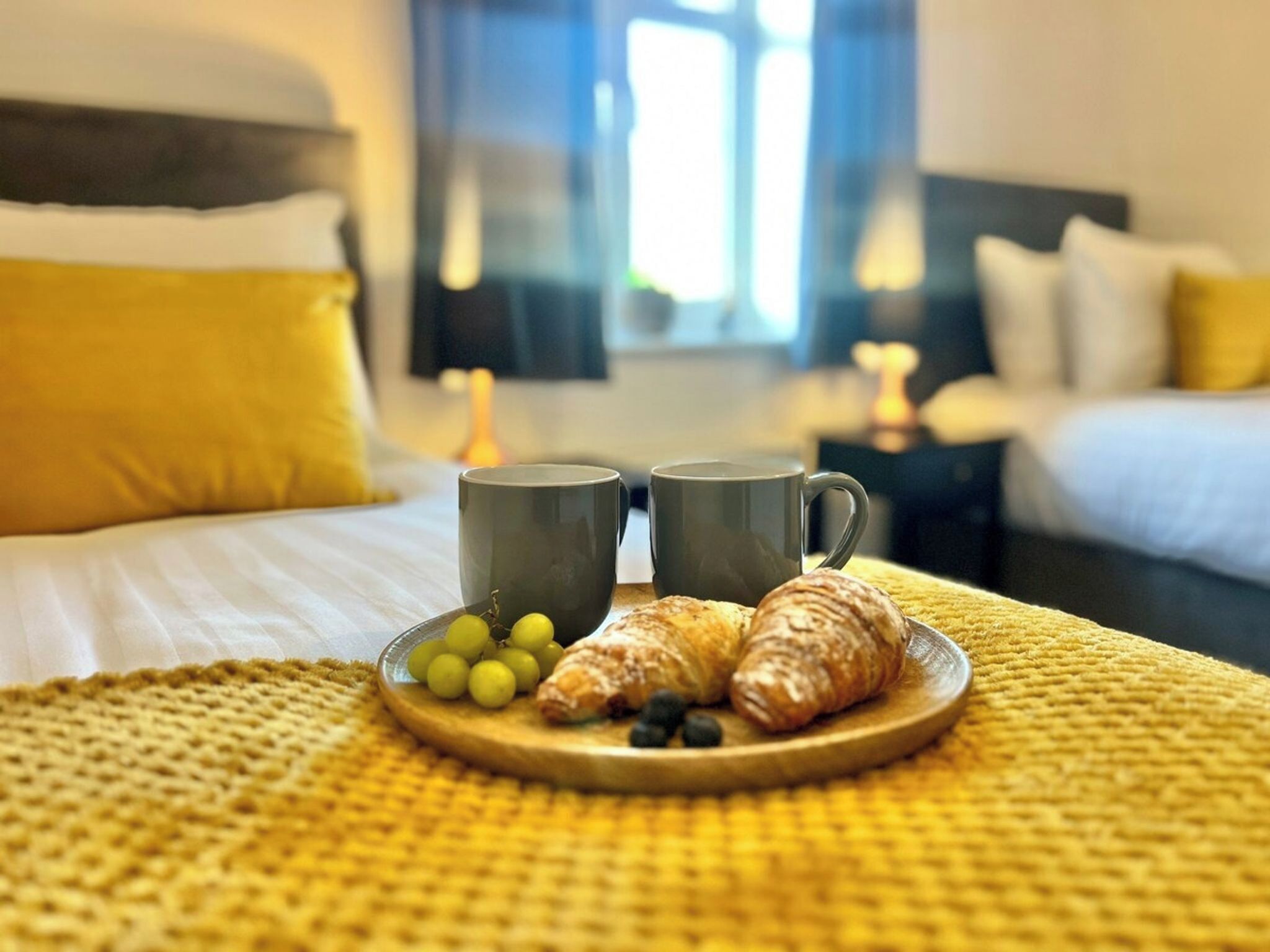 En frukostuppsättning med croissanter och vindruvor på ett fat, två muggar och en säng med gula kuddar.