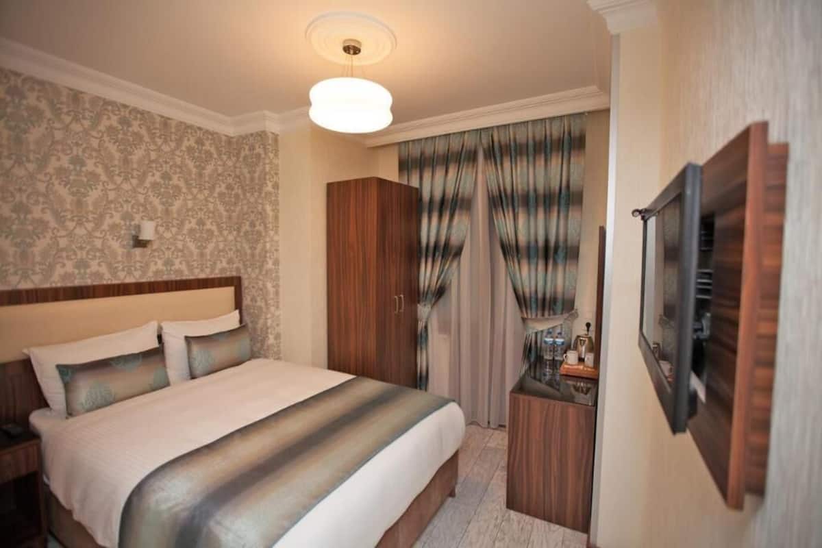 A hotel room with a large bed, a wardrobe, a bedside table with a lamp, and a television mounted on the wall.