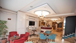 Vestíbulo de hotel con mostrador de recepción, zona de estar con sillas rojas y azules, televisor y candelabro.