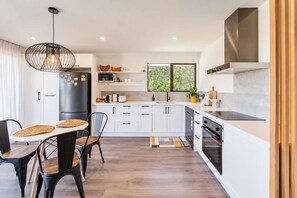 A modern kitchen with white cabinetry, a black refrigerator, and a dining area with a round table and chairs. - Hamptons Cottage | Hot Tub + Fireplace (Prebbleton)