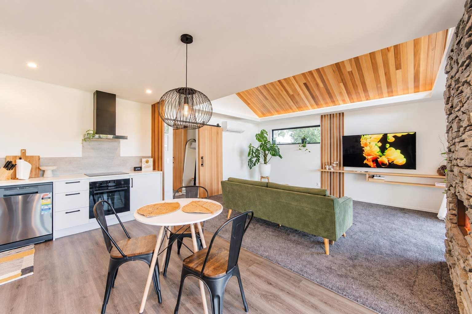 A modern kitchen with a dining area, a green sofa, and a TV on the wall.